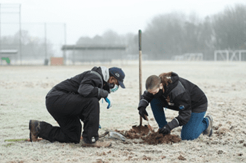 Two Seattle Parks staff, a dark skinned man and a light skinned woman, are kneeled down over a hole they dug investigating the turf at a Seattle Parks athletic field for the newly arrived invasive European Chafer beetle that is damaging grass at parks.