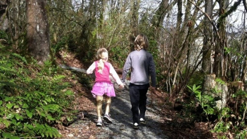 A young boy and girl are walking hand in hand down a gravel path on a sunny day in the woods. 