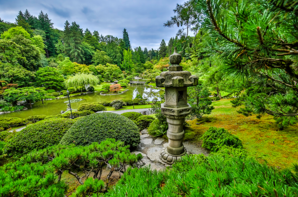 a small pond is surrounded by exquisitely pruned trees of many types, with a stone Japanese sculpture in the foreground