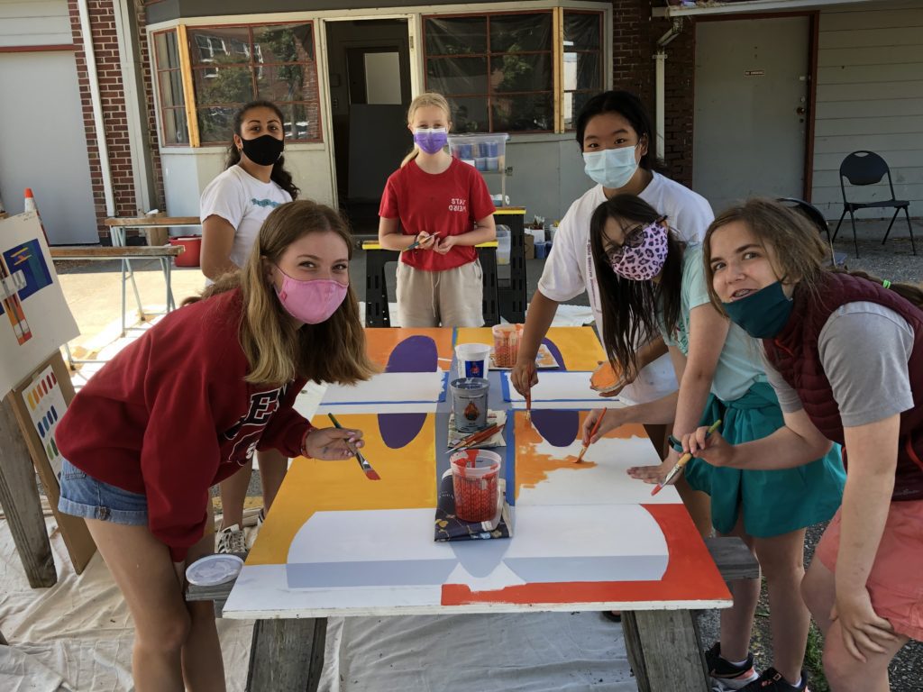 Six teens huddle around a table with paintbrushes in their hands, working on  a large paint-covered board on a table. They wear masks due to the COVID pandemic and are all looking toward the camera.