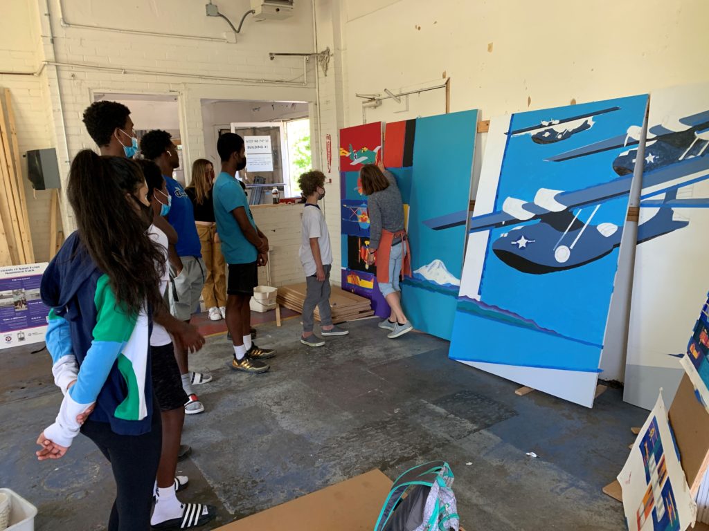 a group of teens stands facing several colorful painted panels while an instructor points out an element on one of the murals
