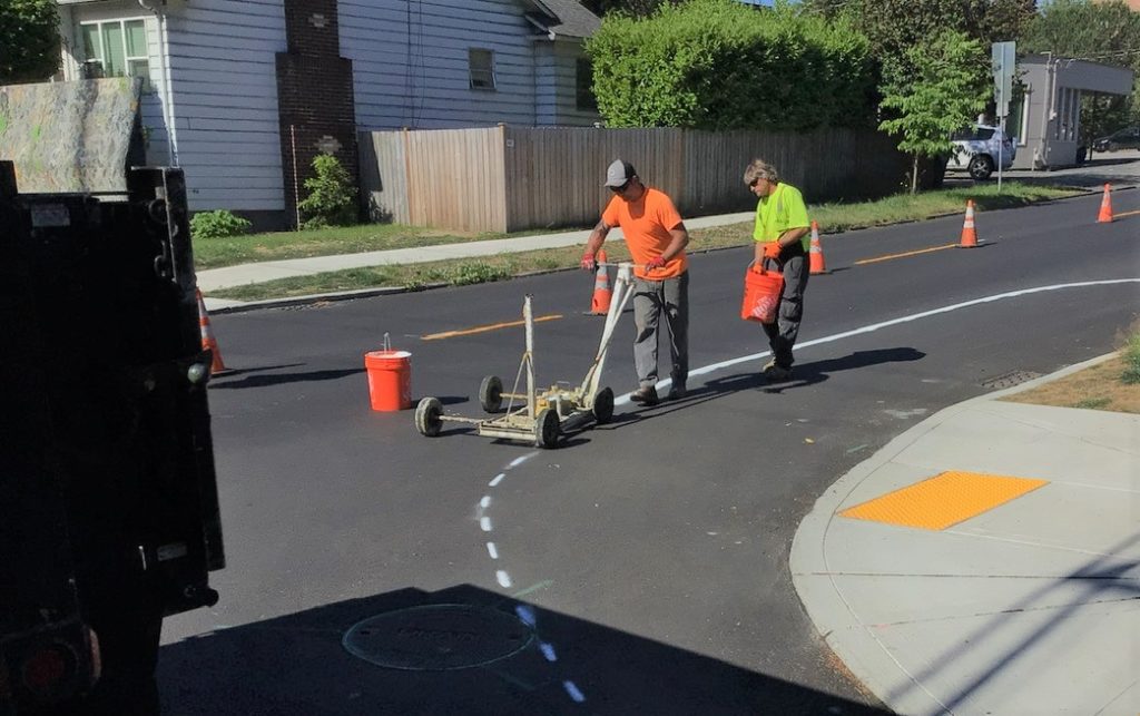 two workers using a wheeled machine to apply paint to the street follow a large truck. Orange safety cones mark the closed part of the street.