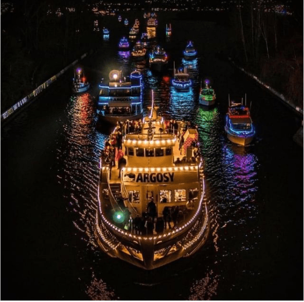 Photo of more than a dozen boats crusing a waterway at night, strung with colorful holiday lights.