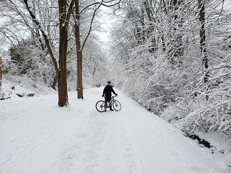 Photo of a bicyclist on a snowy path, surrounded by snow covered trees.  The cyclist is off their bike and facing the camera.