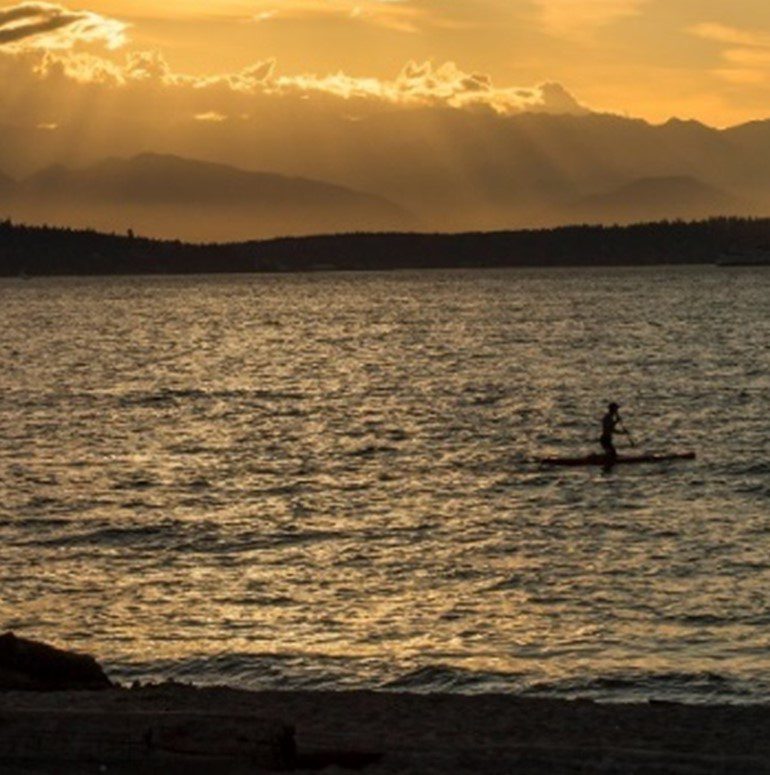 An evening picture looks out onto the Puget Sound with a cloudy yellow sunset. A paddle boarder can be seen in on the saltwater.