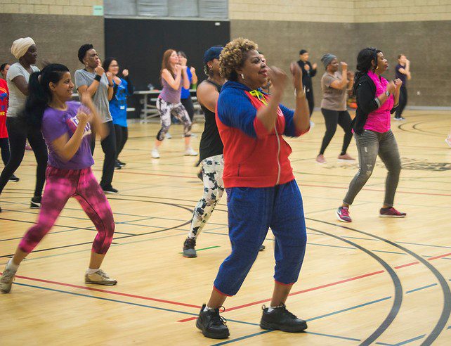 Photo of a dozen or more women in exercise clothing, in a gymnasium, dancing with arms raised and to music and instruction. 