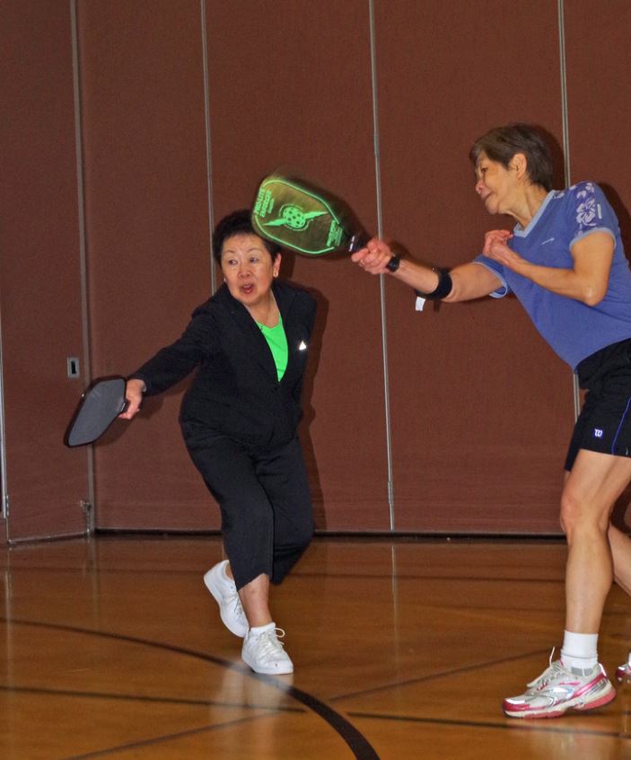 Two people are seen min-swing in a pickleball match in a community center gym. 