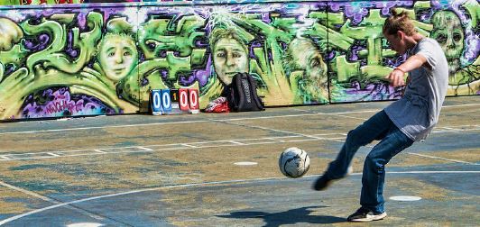 A youth is playing futsal outdoors and is a kicking a soccer ball. The futsal court has a purple and green graffiti art barrier surrounding the court. 
