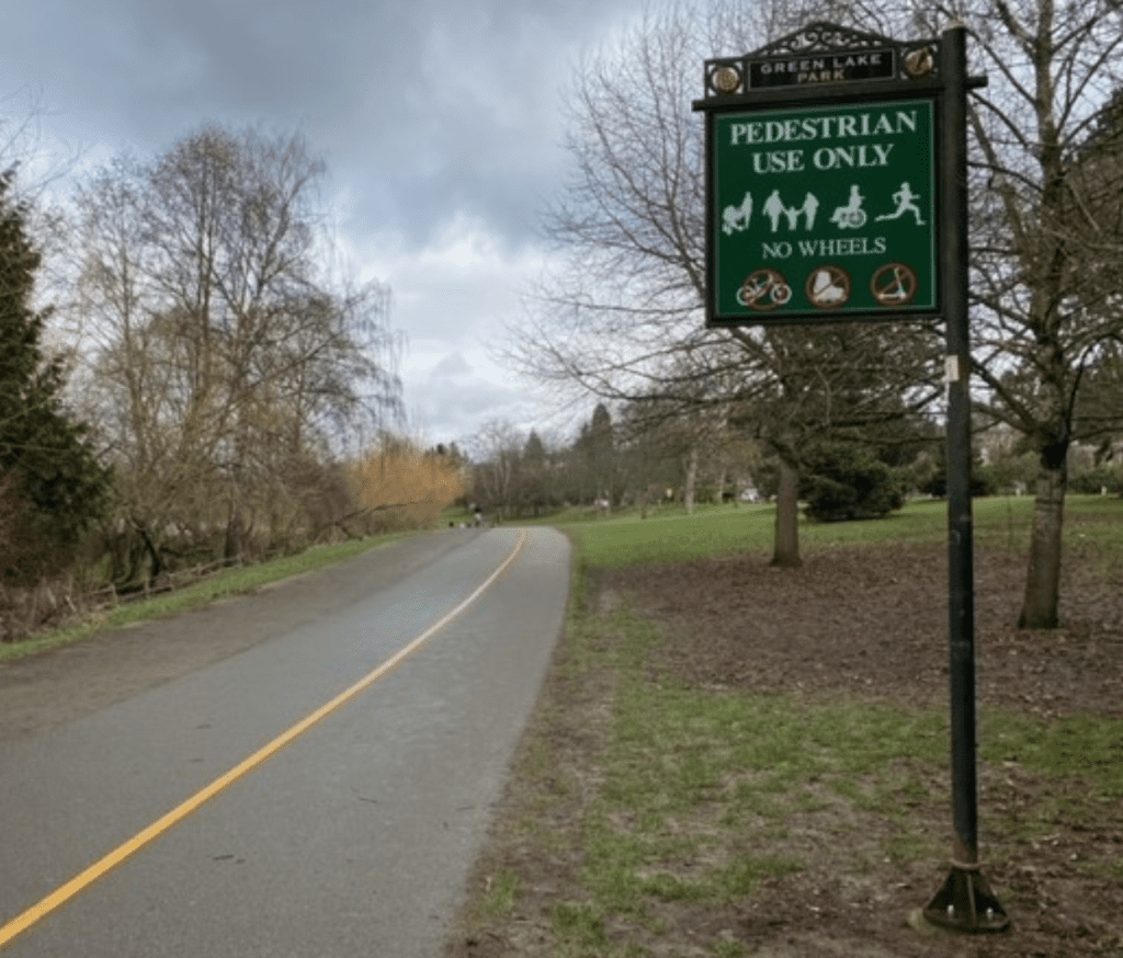 A walking path around Green Lake surrounded by trees and grass. A sign by the side of the trail reads "Pedestrian Use Only, No Wheels"Only
