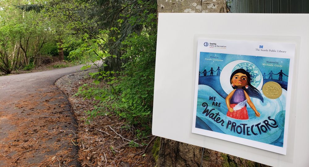 A picture shows a StoryWalk board at a Seattle Park. The Story posted on the board is we Are Water Protectors. A winding path is next to the board. 