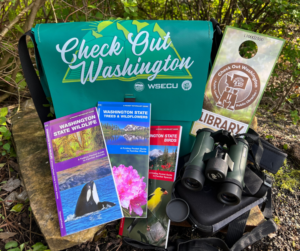 A vareity of maps, a WA State Parks pass, binoculars, and a bag all displayed in a natural outdoor setting