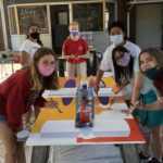 Six teens huddle around a table with paintbrushes in their hands, working on a large paint-covered board on a table. They wear masks due to the COVID pandemic and are all looking toward the camera.