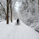 Photo of a bicyclist on a snowy path, surrounded by snow covered trees. The cyclist is off their bike and facing the camera.