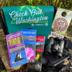 A vareity of maps, a WA State Parks pass, binoculars, and a bag all displayed in a natural outdoor setting