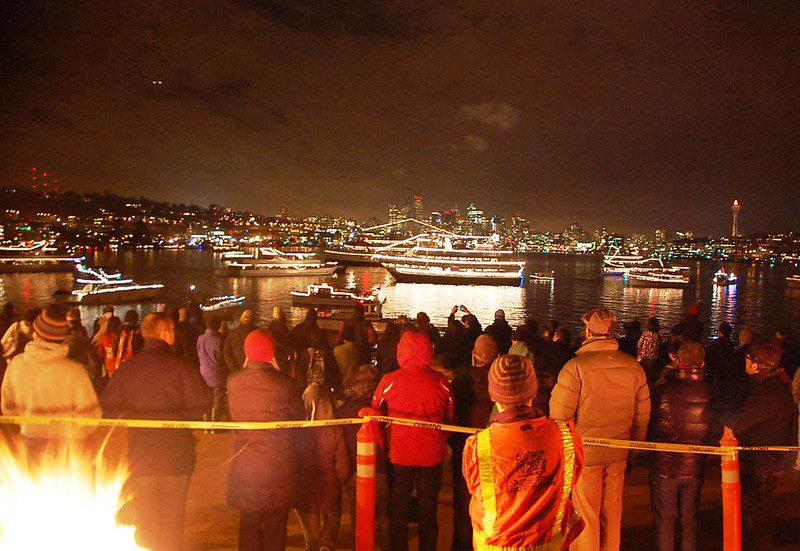 People gathered outdoors at night around a bonfire watching ships in the lake