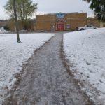 Brick building int he background with a snow covered lawn and path in the foreground