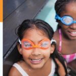 Two children in a pool smiling at the camera while wearing swim goggles.