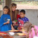 A teacher with children outdoors holding a leaf during a lesson. Students are looking at the leaf and others are writing in a notebook.