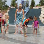 Adults and children stand in a splashpad outdoors on a sunny day with water splashing at their feet.