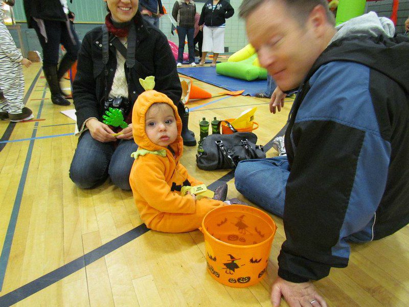 2 parents accompany a baby in a pumpkin costume in a gymnasium.  To the front, an empty orange candy bucket.  Behind them is a slide on a mat.