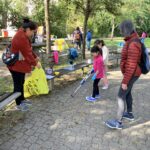 A child uses a litter picker to collect trash in a park while adults and other children participate in a community cleanup event.