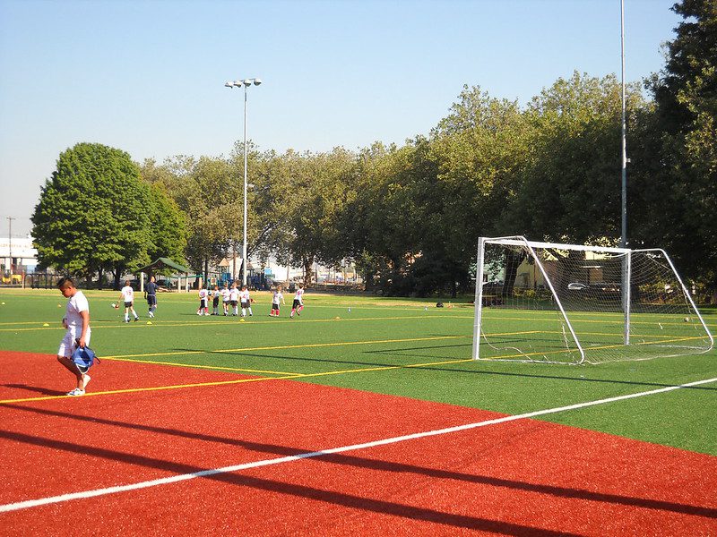 A synthetic field with soccer goal and children playing a game of soccer under a blue sky surrounded by trees