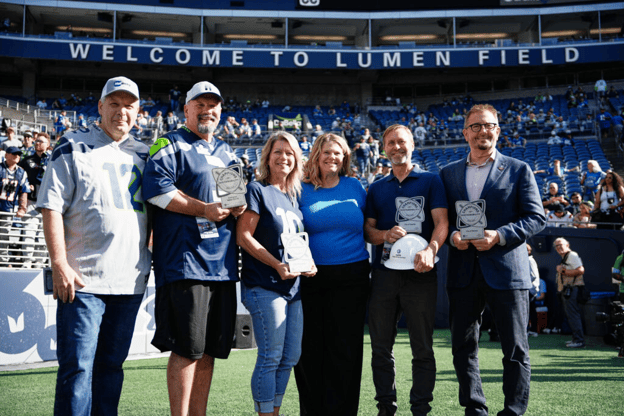 Six people on the field of an athletic stadium pose for the camera, with spectators filling the seats behind them.